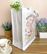A side view white canvas tote bag with a gossipy llama surrounded by flowers in a vintage-style logo for "Drama Llama Bleater&