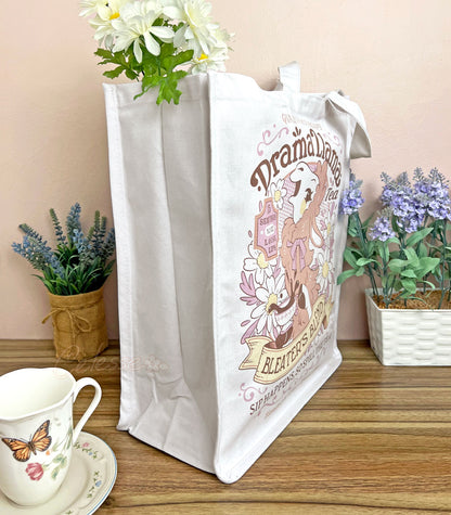 A side view white canvas tote bag with a gossipy llama surrounded by flowers in a vintage-style logo for "Drama Llama Bleater&