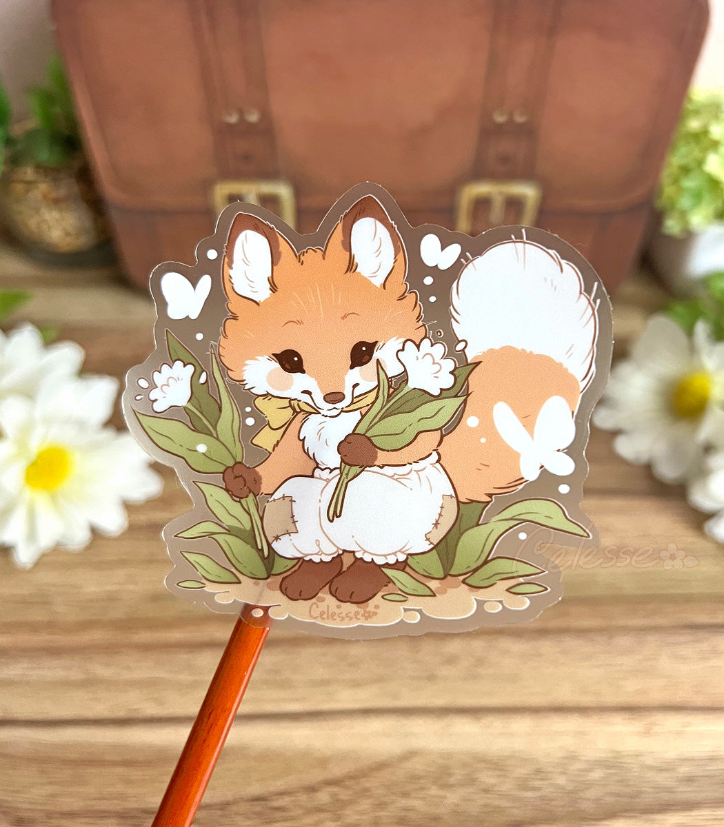 A photo of a clear vinyl sticker with artwork of a baby fox with flowers above a wooden surface with a blurred background.