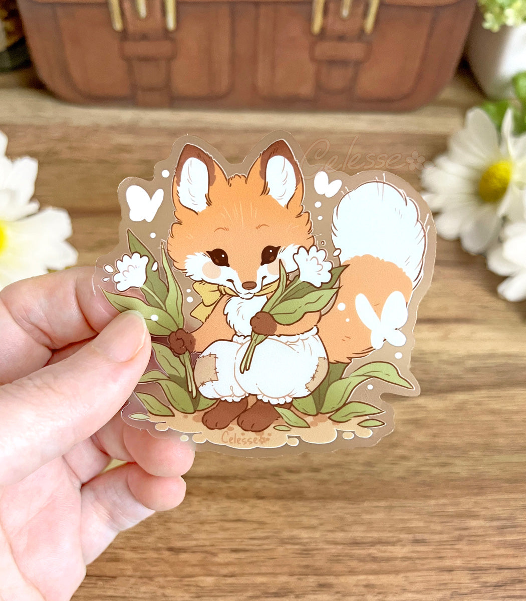 A photo of a hand holding a clear vinyl sticker with artwork of a baby fox with flowers above a wooden surface with a blurred background.