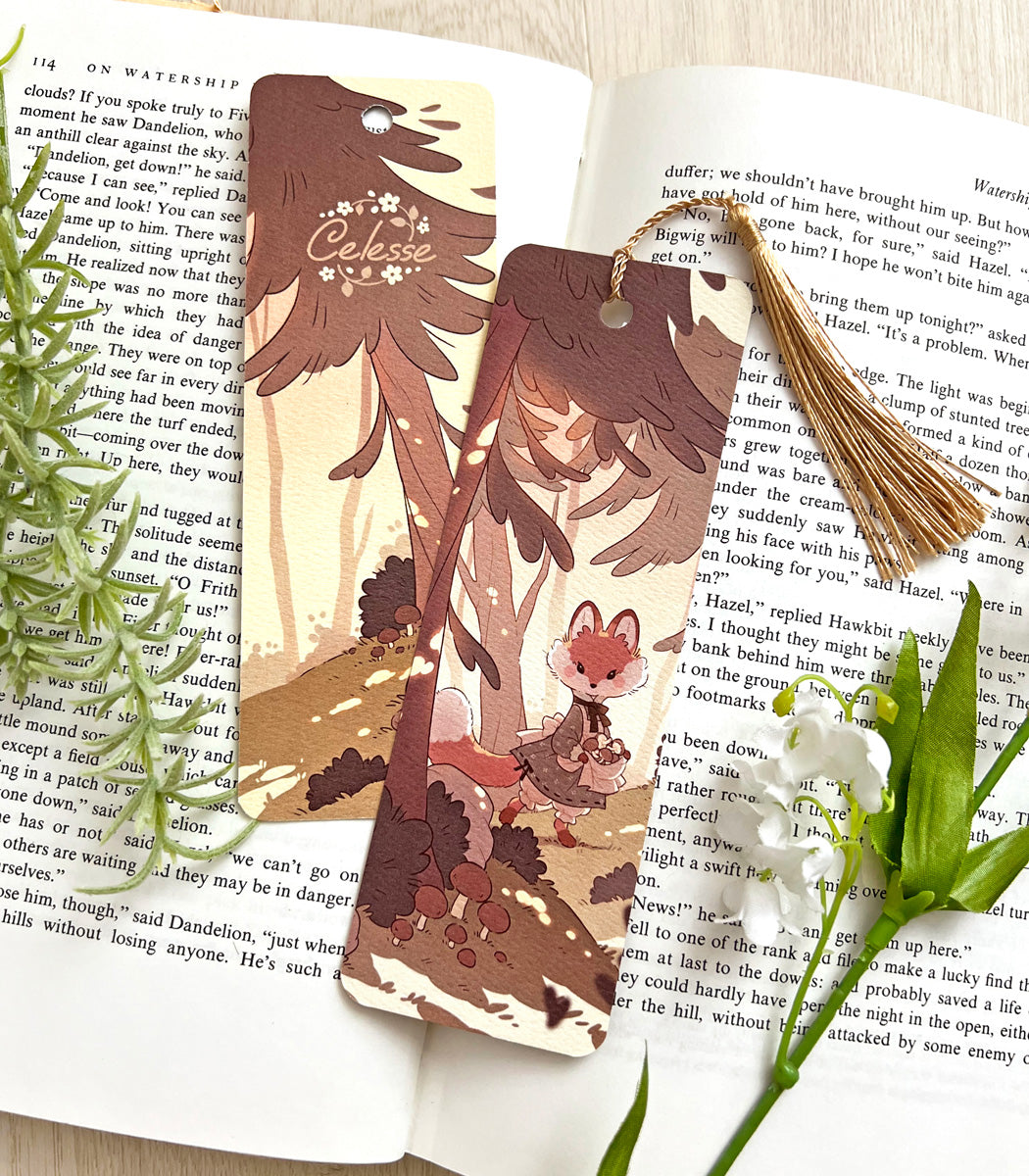 A photo of a bookmark placed atop an open book, the artwork featuring a fox wearing a vintage-style dress while roaming the forest in the warm glow of evening, picking mushrooms.
