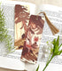 A photo of a bookmark placed atop an open book, the artwork featuring a fox wearing a vintage-style dress while roaming the forest in the warm glow of evening, picking mushrooms.