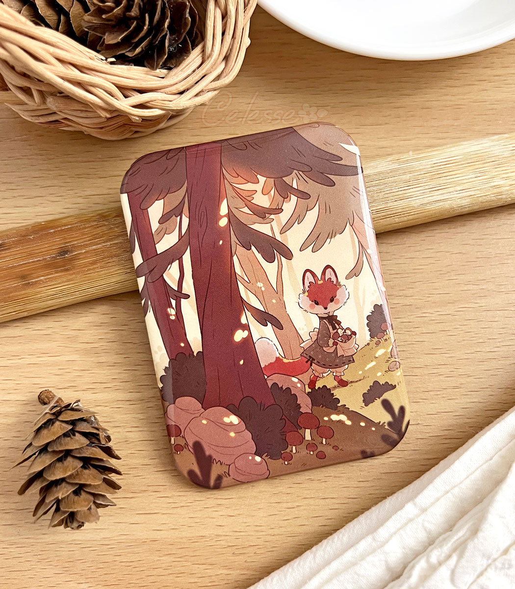 A photo of a fridge magnet laid on a table with artwork featuring a fox in a simple dress walking through a sunlit forest and collecting mushrooms