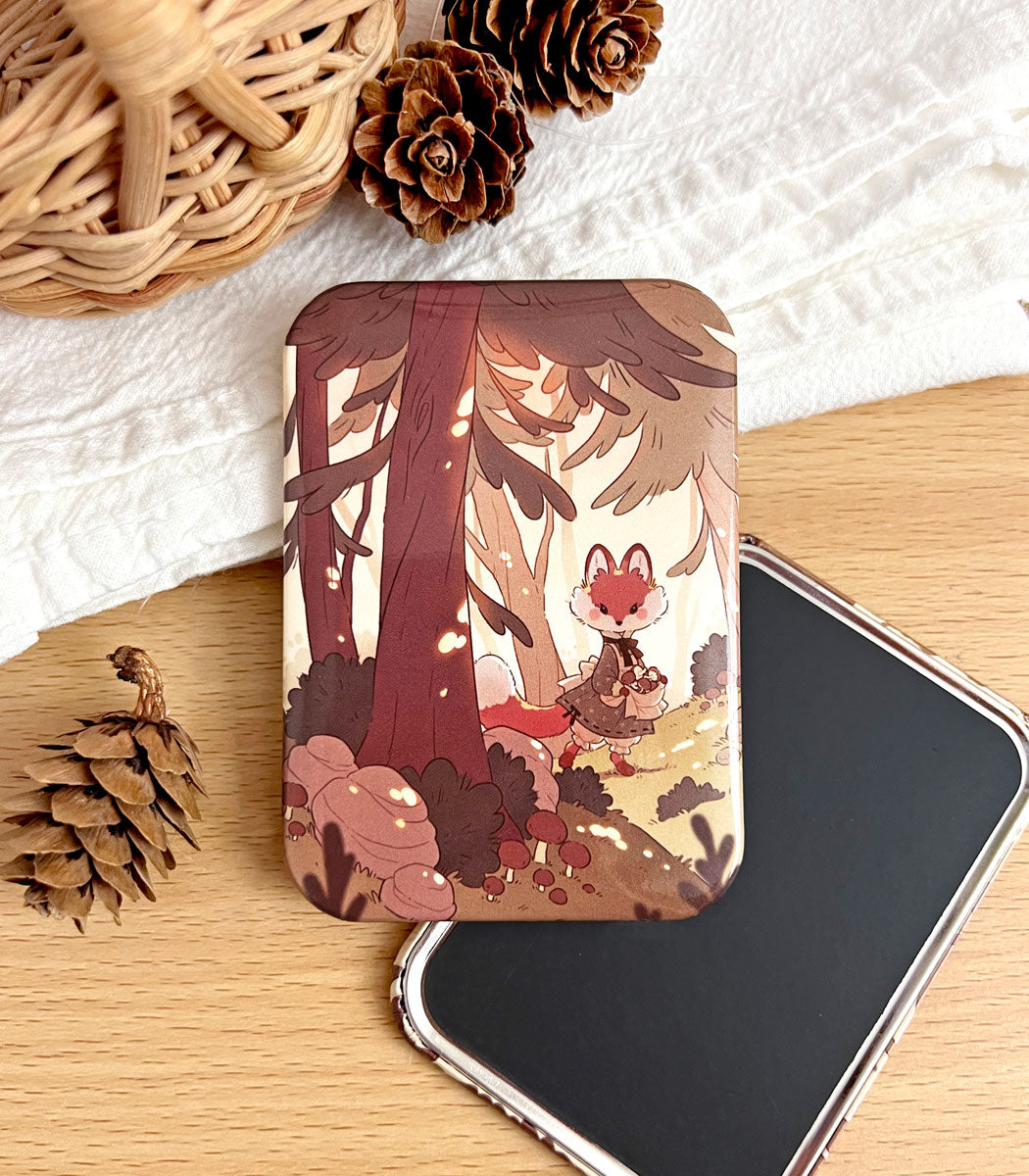 A photo of a fridge magnet laid on a table with artwork featuring a fox in a simple dress walking through a sunlit forest and collecting mushrooms