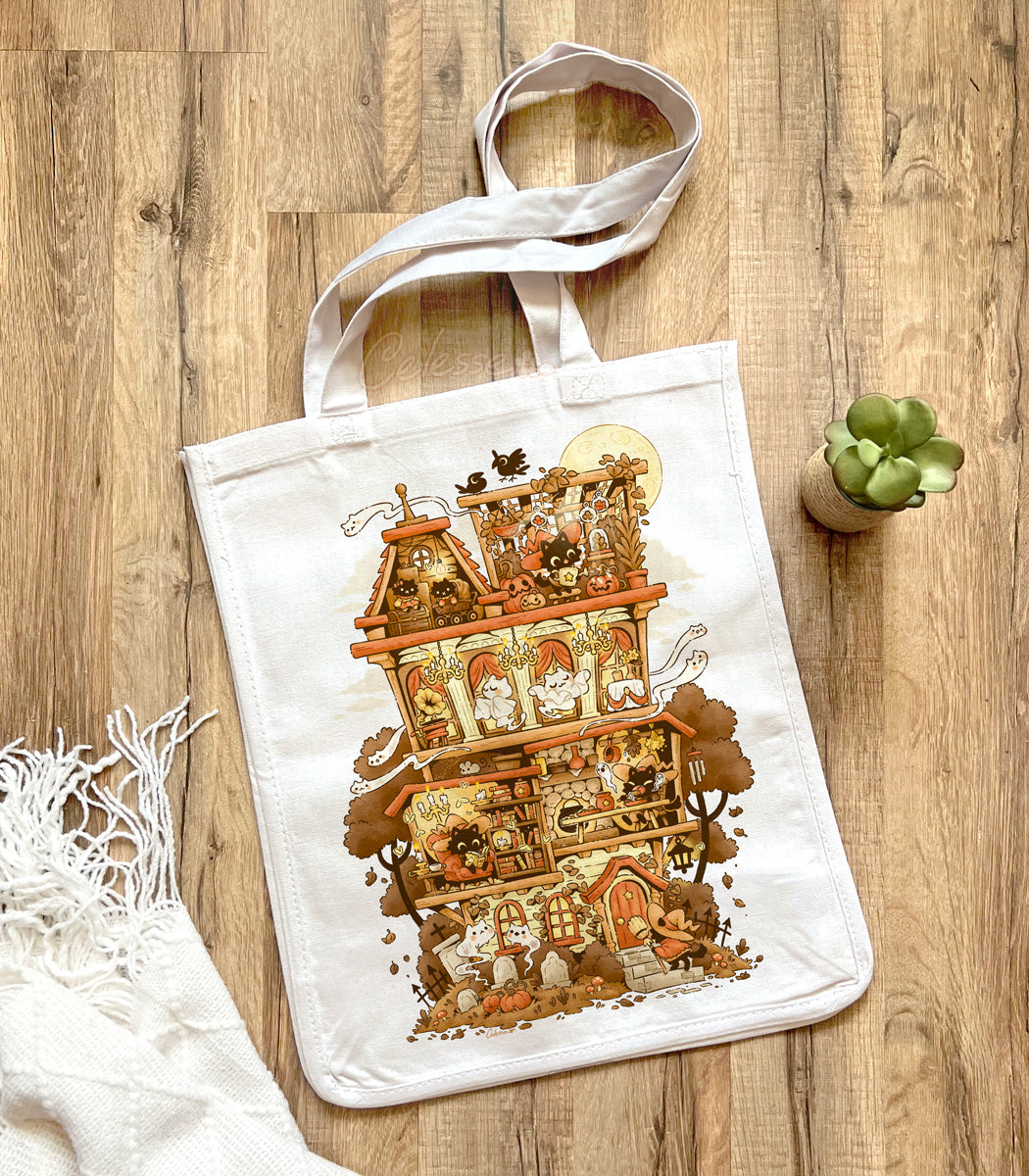 White tote bag with a printed design featuring a Halloween-themed haunted home full of witch cats and ghosts, laid out on a wooden surface.