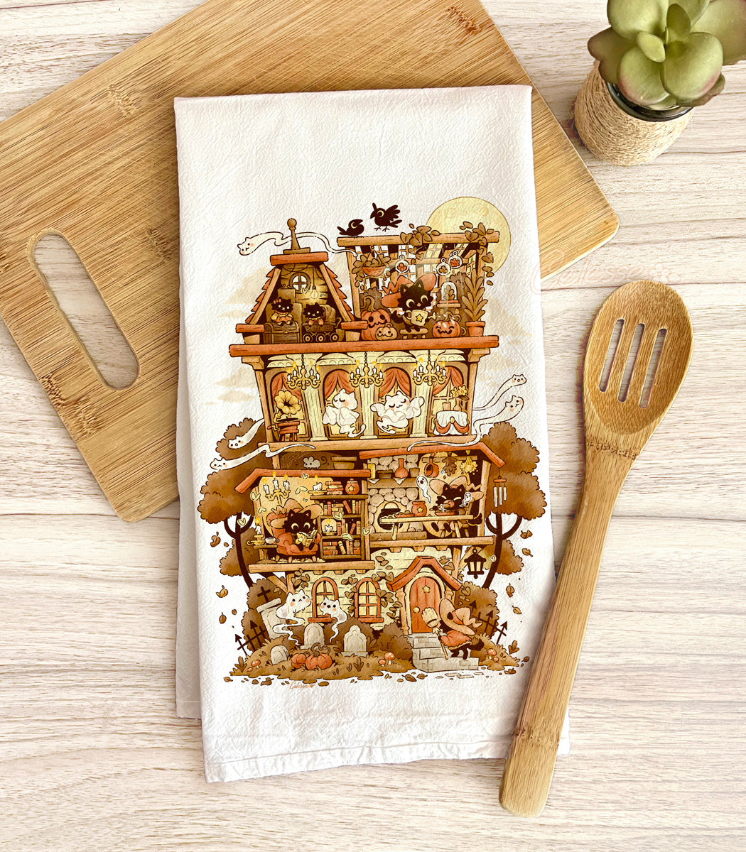 A flour sack tea towel with a detailed illustration of a Halloween-themed haunted home full of witch cats and ghosts, laid out on a wooden surface with a spoon and plant.