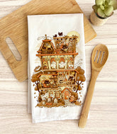 A flour sack tea towel with a detailed illustration of a Halloween-themed haunted home full of witch cats and ghosts, laid out on a wooden surface with a spoon and plant.