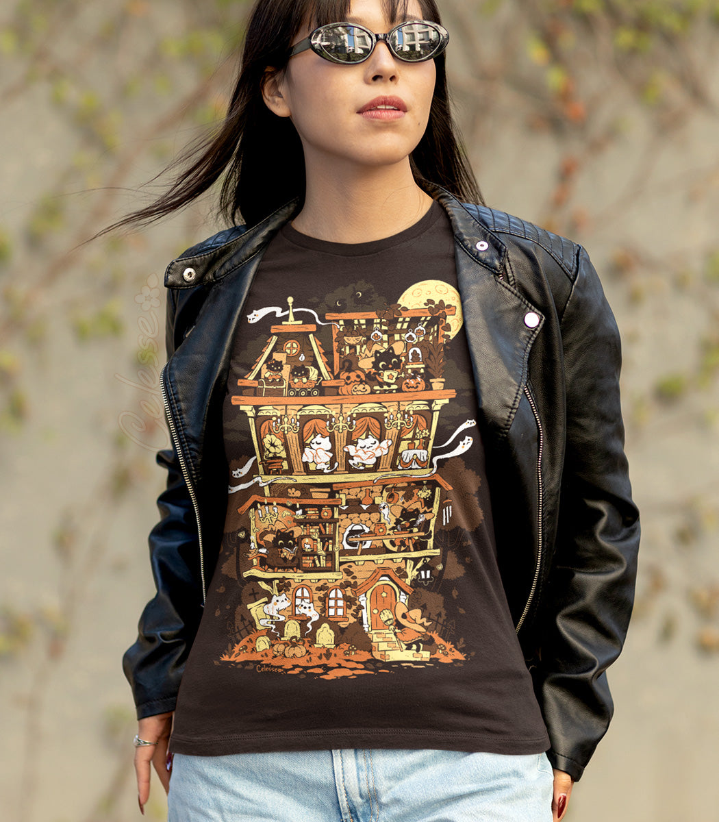A photo of a person standing outdoors in a jacket and sunglasses, wearing a  dark brown shirt with the print featuring a Halloween-themed haunted home full of witch cats and ghosts.