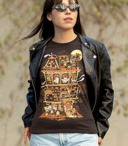 Person standing outdoors in a jacket and sunglasses, wearing a  dark brown shirt with the print featuring a Halloween-themed haunted home full of witch cats and ghosts.