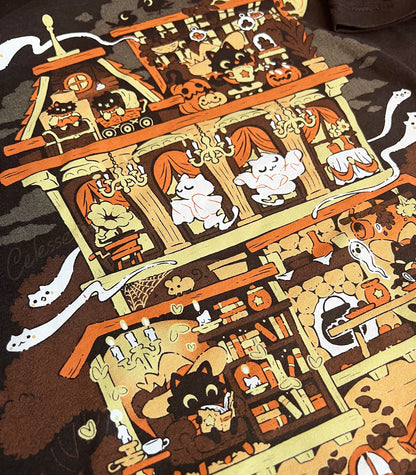 A close-up photo of a dark brown shirt, the print featuring a whimsical Halloween-themed haunted home full of witch cats and ghosts.