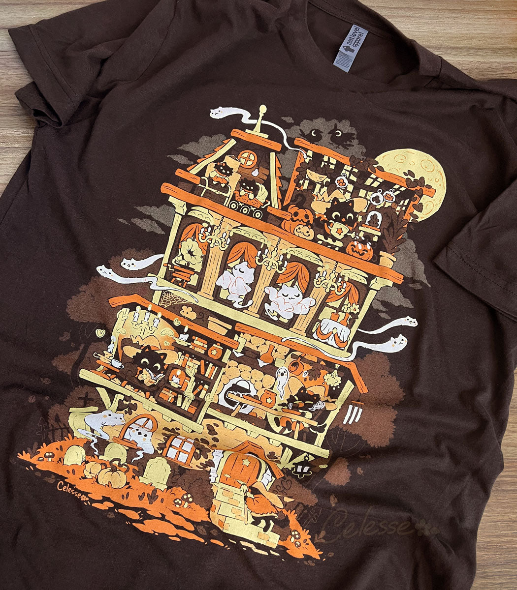A photo of a dark brown shirt laid on a wooden table, the print featuring a whimsical Halloween-themed haunted home full of witch cats and ghosts.