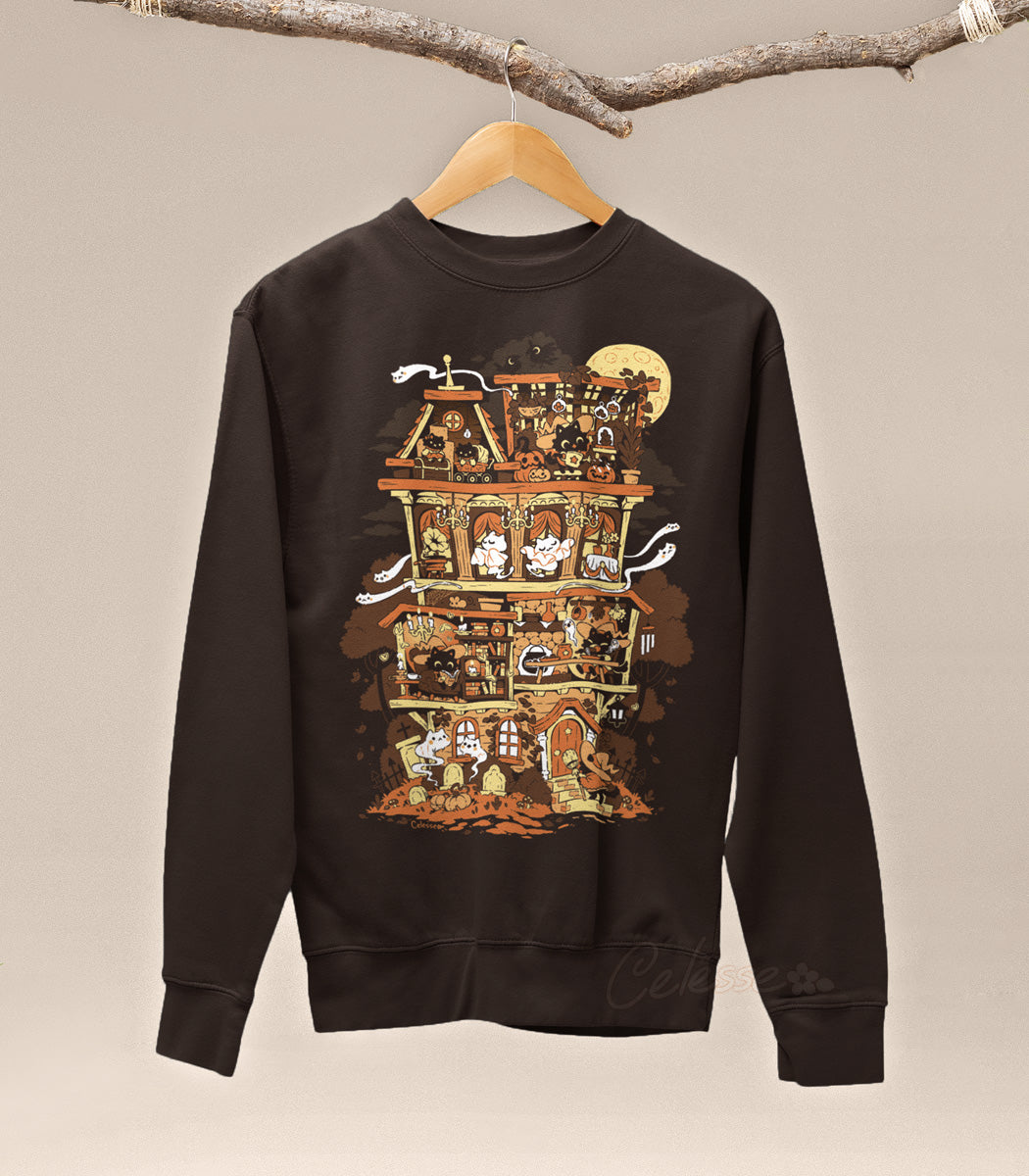 A dark brown sweatshirt on a wood hanger hung from a branch, the print featuring a Halloween-themed haunted home full of witch cats and ghosts.