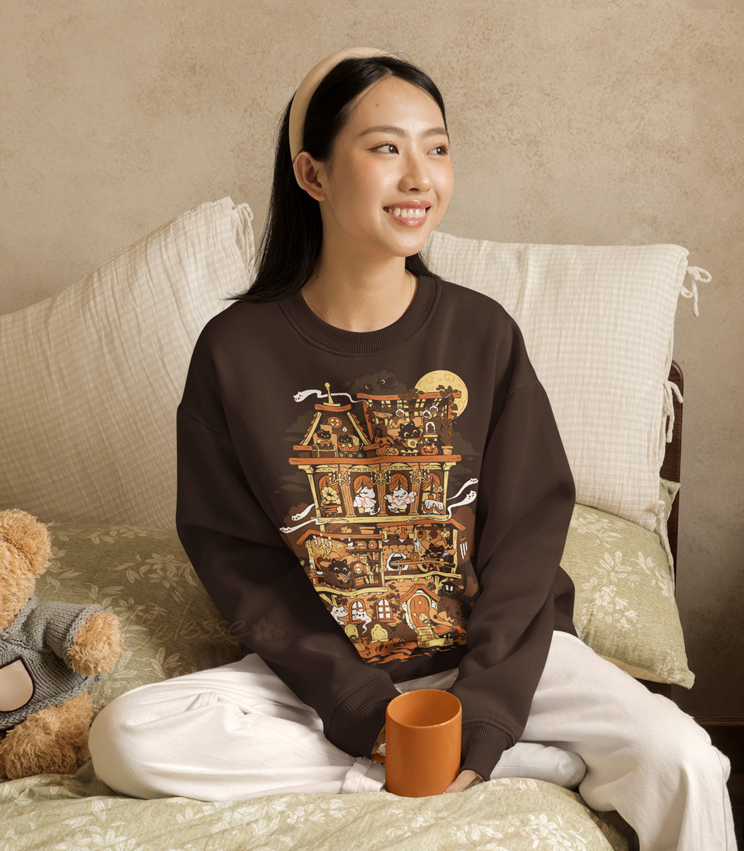 Woman wearing a brown sweatshirt with a Halloween-themed print of a haunted home full of witch cats, sitting on a bed with a teddy bear and mug.