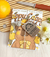 A folded kitchen tea towel tied with twine with a vintage-style logo for Happy Cappy Yuzu Fruit featuring a blissful capybara, styled on a wooden cutting board next to an orange, a small knife, and white flowers.