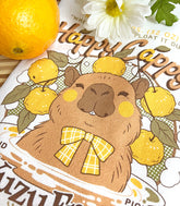 A close up of a white kitchen tea towel with a vintage-style logo for Happy Cappy Yuzu Fruit featuring a blissful capybara, styled on a wooden cutting board next to an orange and white flowers.