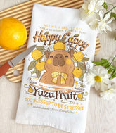 A white kitchen tea towel with a vintage-style logo for Happy Cappy Yuzu Fruit featuring a blissful capybara, styled on a wooden cutting board next to an orange, a small knife, and white flowers.