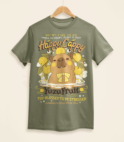 A light olive green shirt on a wood hanger with a print of a vintage-style logo for Happy Cappy Yuzu Fruit, the center art featuring a smiling capybara in a hot spring.