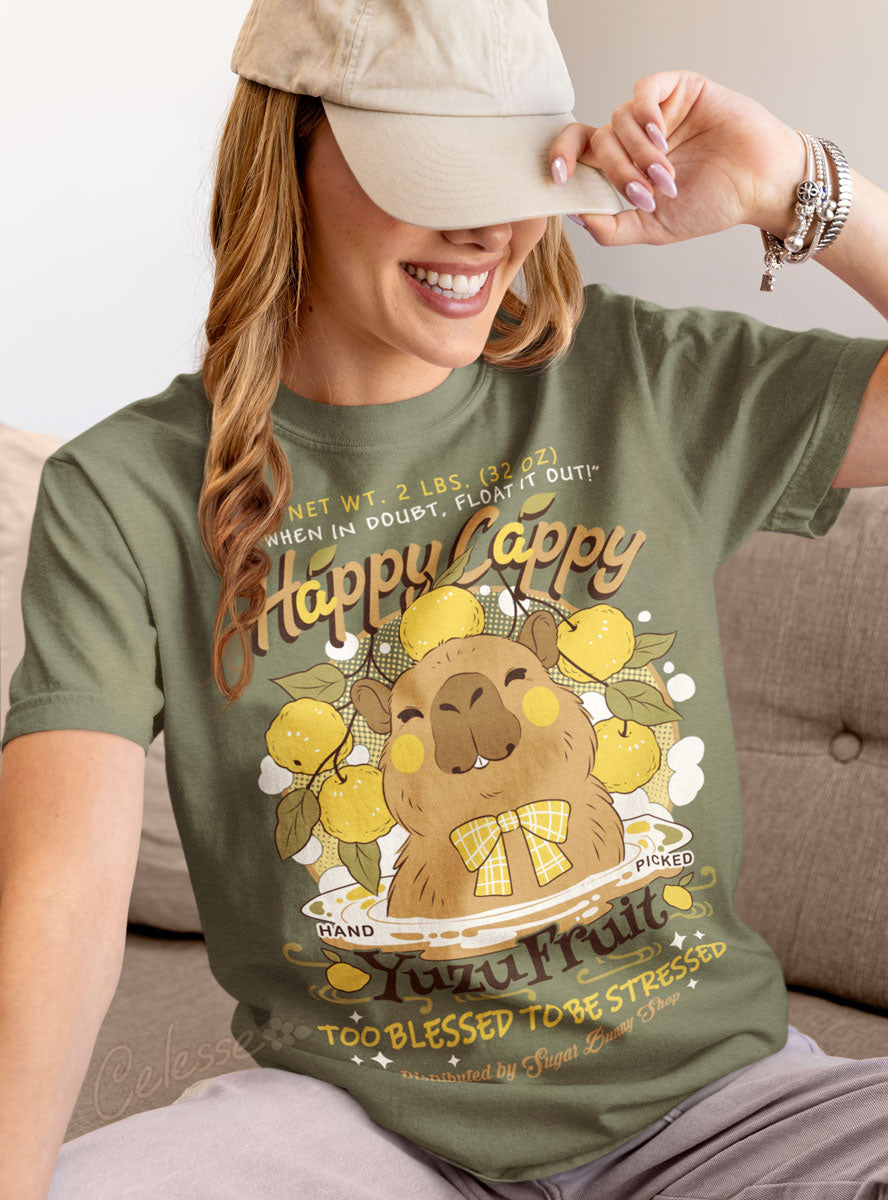 A woman sitting on a couch and pulling a baseball cap down over her eyes, wearing a light olive green shirt with a print of a vintage-style logo for Happy Cappy Yuzu Fruit, the center art featuring a smiling capybara in a hot spring. 