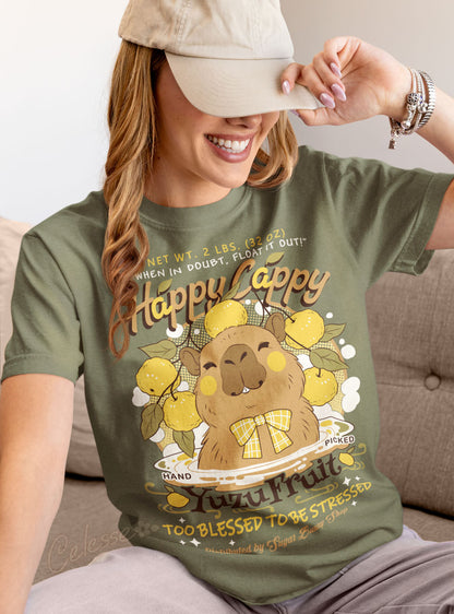 A woman sitting on a couch and pulling a baseball cap down over her eyes, wearing a light olive green shirt with a print of a vintage-style logo for Happy Cappy Yuzu Fruit, the center art featuring a smiling capybara in a hot spring. 