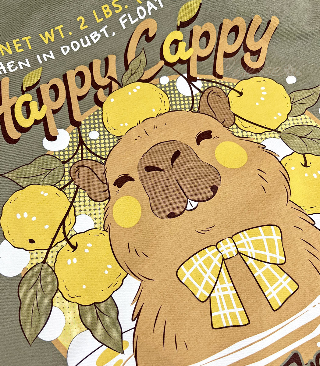 A close up of a light olive green shirt with a print of a vintage-style logo for Happy Cappy Yuzu Fruit, the center art featuring a smiling capybara in a hot spring.