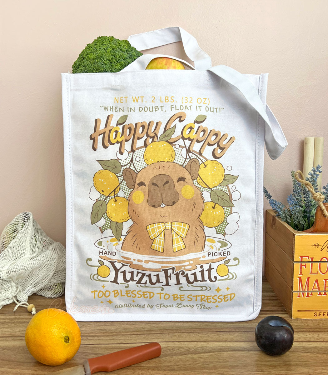 A white canvas tote bag with a cute capybara in a logo for "Happy Cappy Yuzu Fruit," styled on a wood table with produce, an orange, a knife, and a small wooden box in the background.