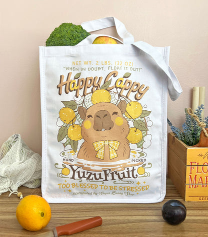 A white canvas tote bag with a cute capybara in a logo for "Happy Cappy Yuzu Fruit," styled on a wood table with produce, an orange, a knife, and a small wooden box in the background.