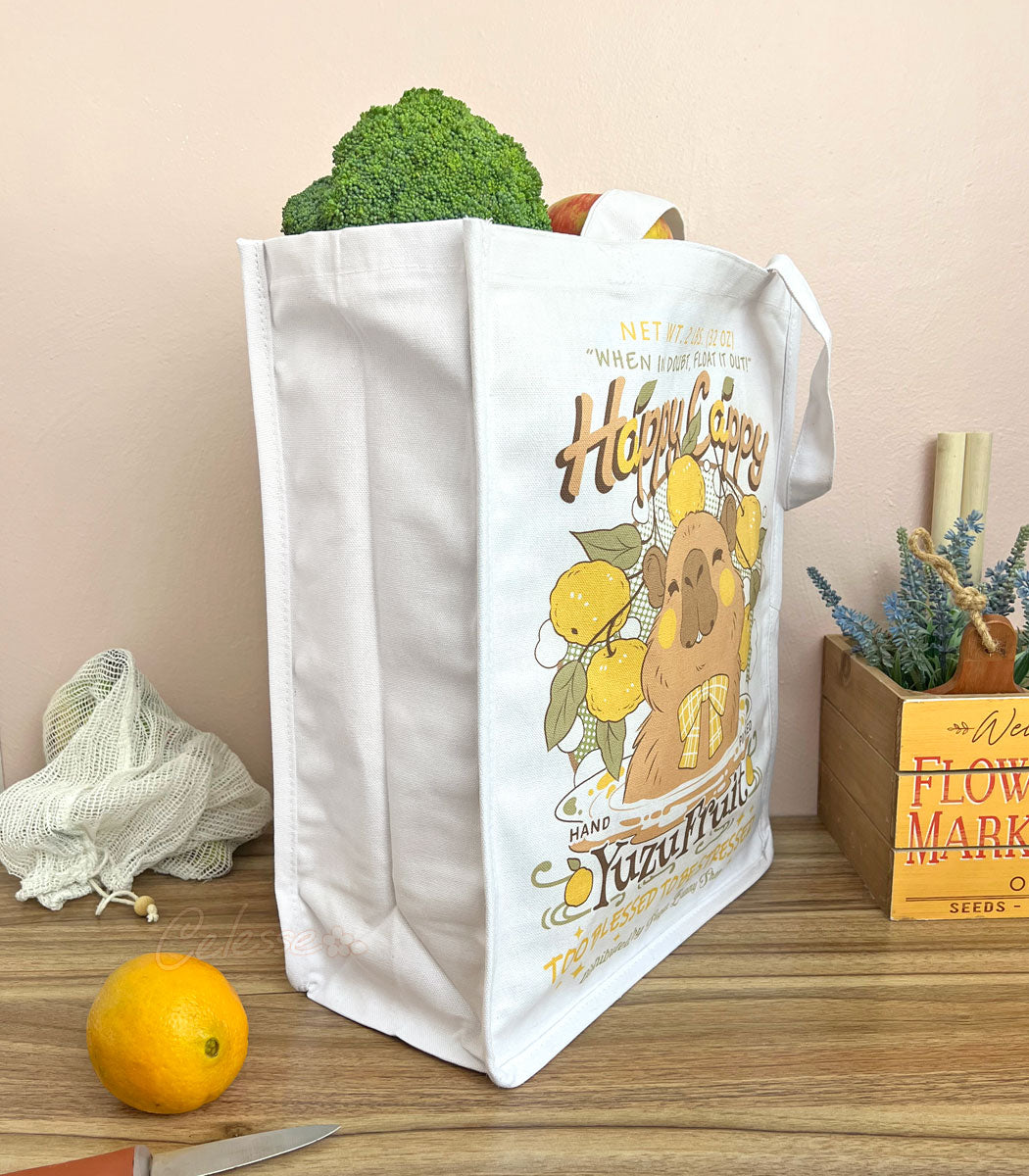 A side view of a white canvas tote bag with a cute capybara in a logo for "Happy Cappy Yuzu Fruit," styled on a wood table with produce, an orange, a knife, and a small wooden box in the background.