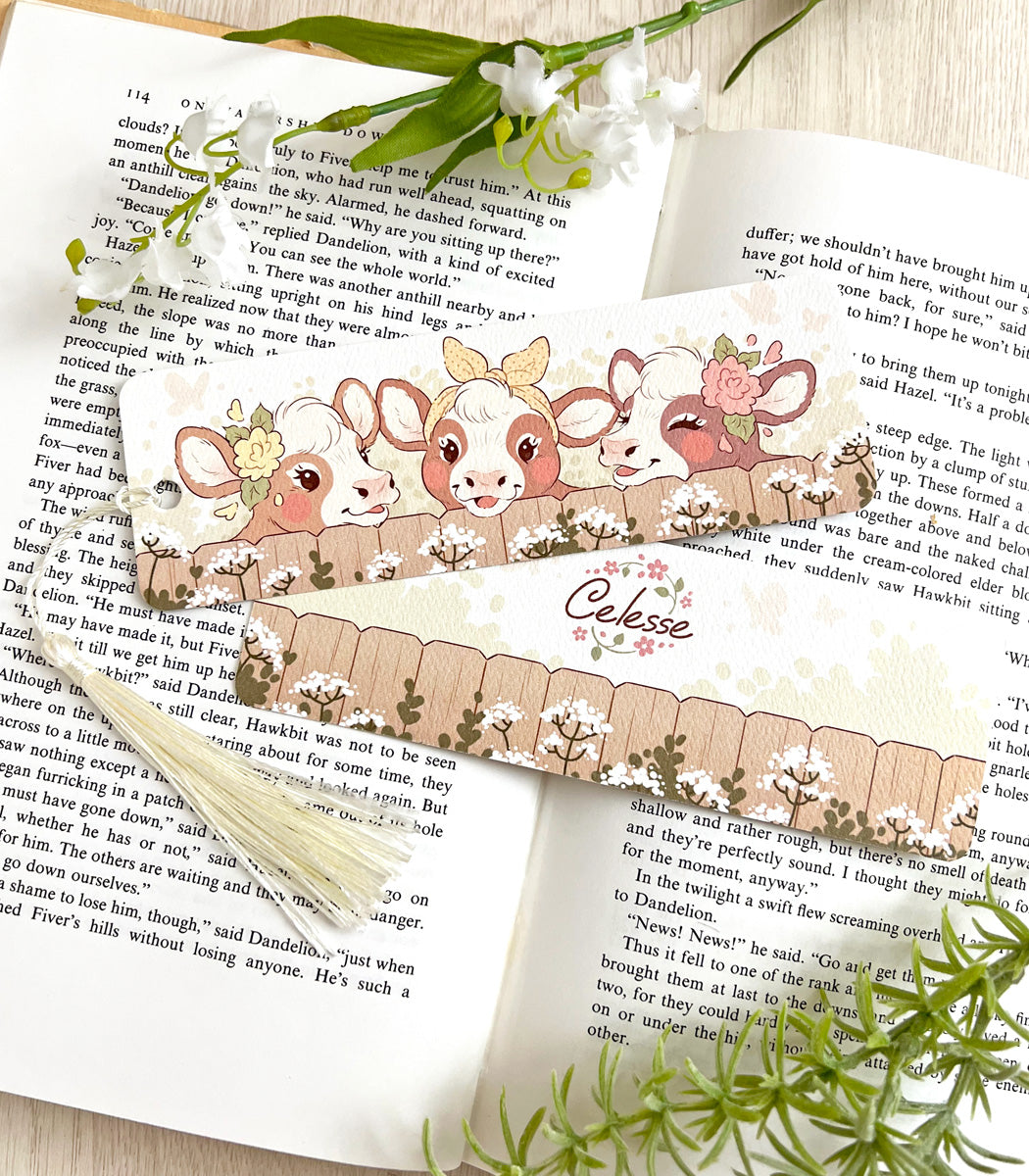 A photo of a bookmark placed atop an open book, the artwork featuring three country cows peeking over the top of a fence.