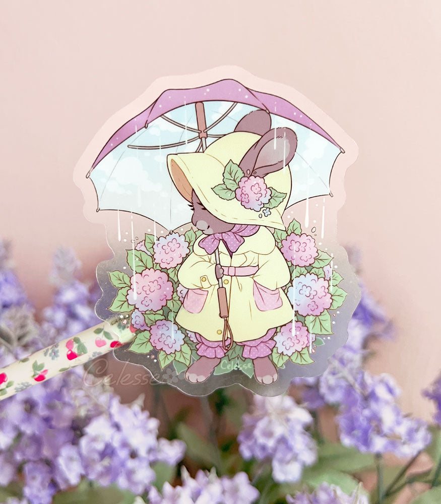 A photo of a clear vinyl sticker featuring artwork of a rabbit in a vintage style raincoat and bonnet, standing among hydrangea flowers and holding an umbrella as rain sprinkles down.