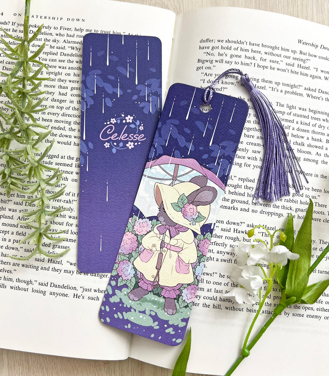 A photo of a bookmark placed atop an open book, the artwork featuring a bunny in an old-fashioned raincoat, holding an umbrella under the rain while standing among hydrangea bushes.