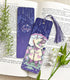 A photo of a bookmark placed atop an open book, the artwork featuring a bunny in an old-fashioned raincoat, holding an umbrella under the rain while standing among hydrangea bushes.