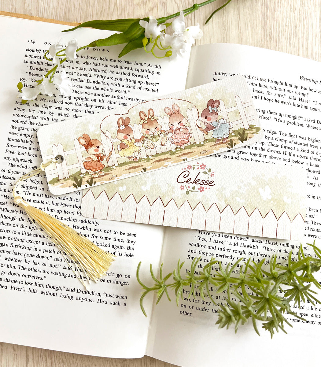 A photo of a bookmark placed atop an open book, the artwork featuring a group of young bunny girls in vintage dresses playing jump rope together.