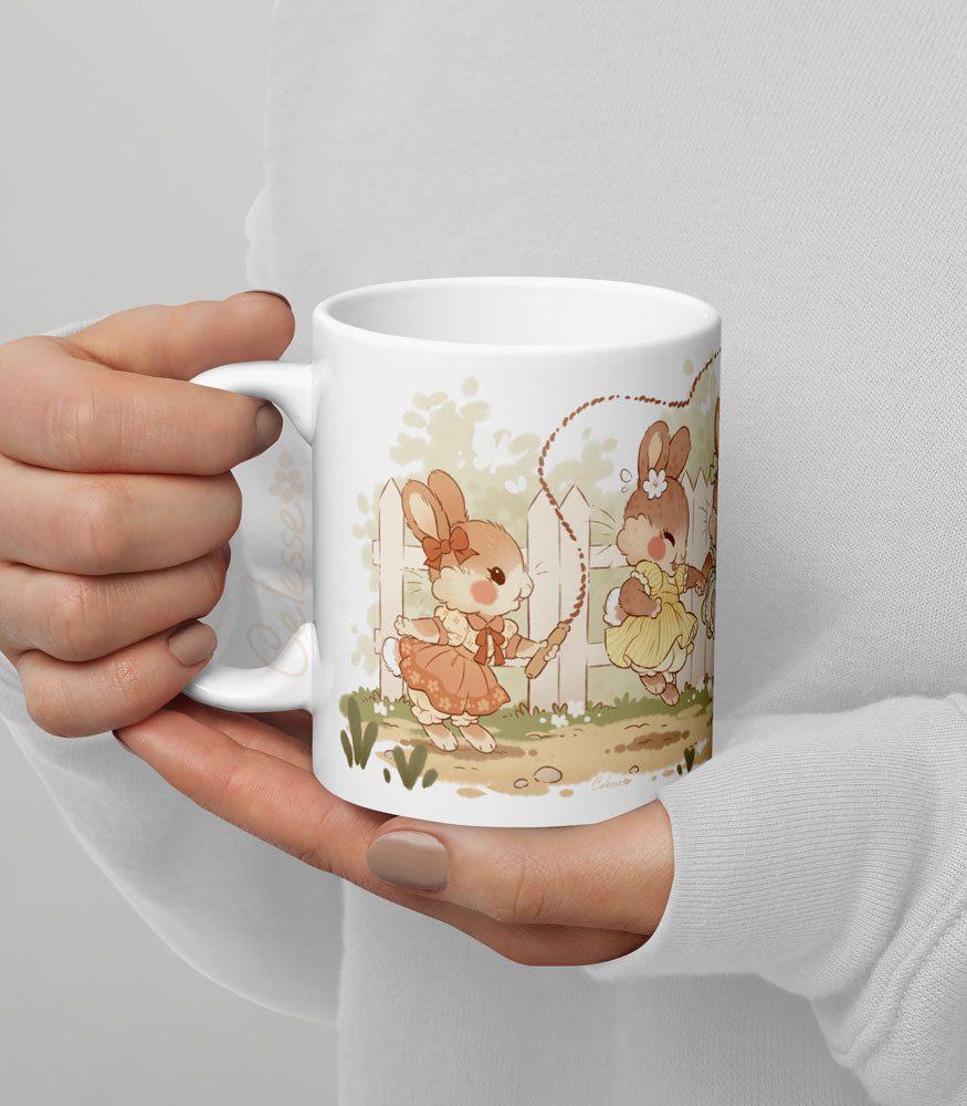 Hands holding an 11 oz. ceramic mug with a print featuring art of a bunch of bunny girls playing jump rope in vintage dresses.