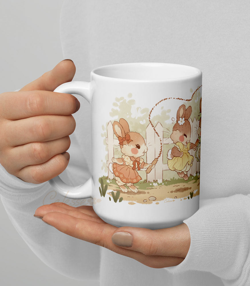 Hands holding an 15 oz. ceramic mug with a print featuring art of a bunch of bunny girls playing jump rope in vintage dresses.