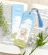 A photo of a bookmark placed atop an open book, the artwork featuring a bunny in a vintage dress and bonnet perched serenely atop a grassy hill on a sunny day while a breeze blows. The text reads "Just be..."