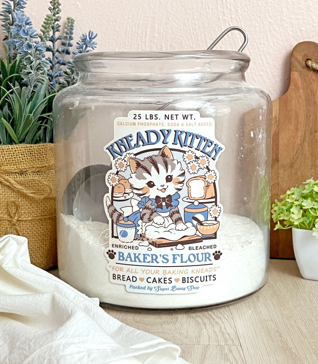 A photo of a large glass flour container on a counter with plants, a cutting board and a tea towel surrounding it. The container has a label sticker on it featuring a faux logo for Kneady Kitten Baker&