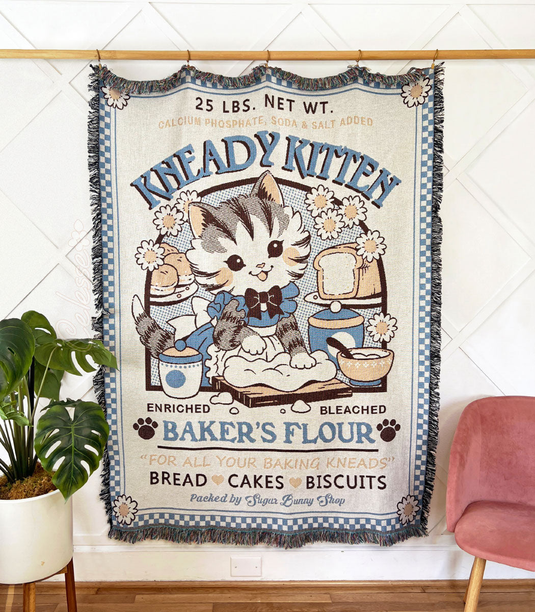 A photo of a woven tapestry blanket hung from a wooden pole against a wall, a plant and chair on either side of it. The blanket features a vintage-style logo for Kneady Kitten Baker&