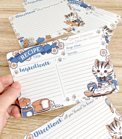 A hand holding a recipe card over a wooden surface with more cards scattered around, the design featuring vintage-style art of a baking kitten wearing a frilly dress and apron.