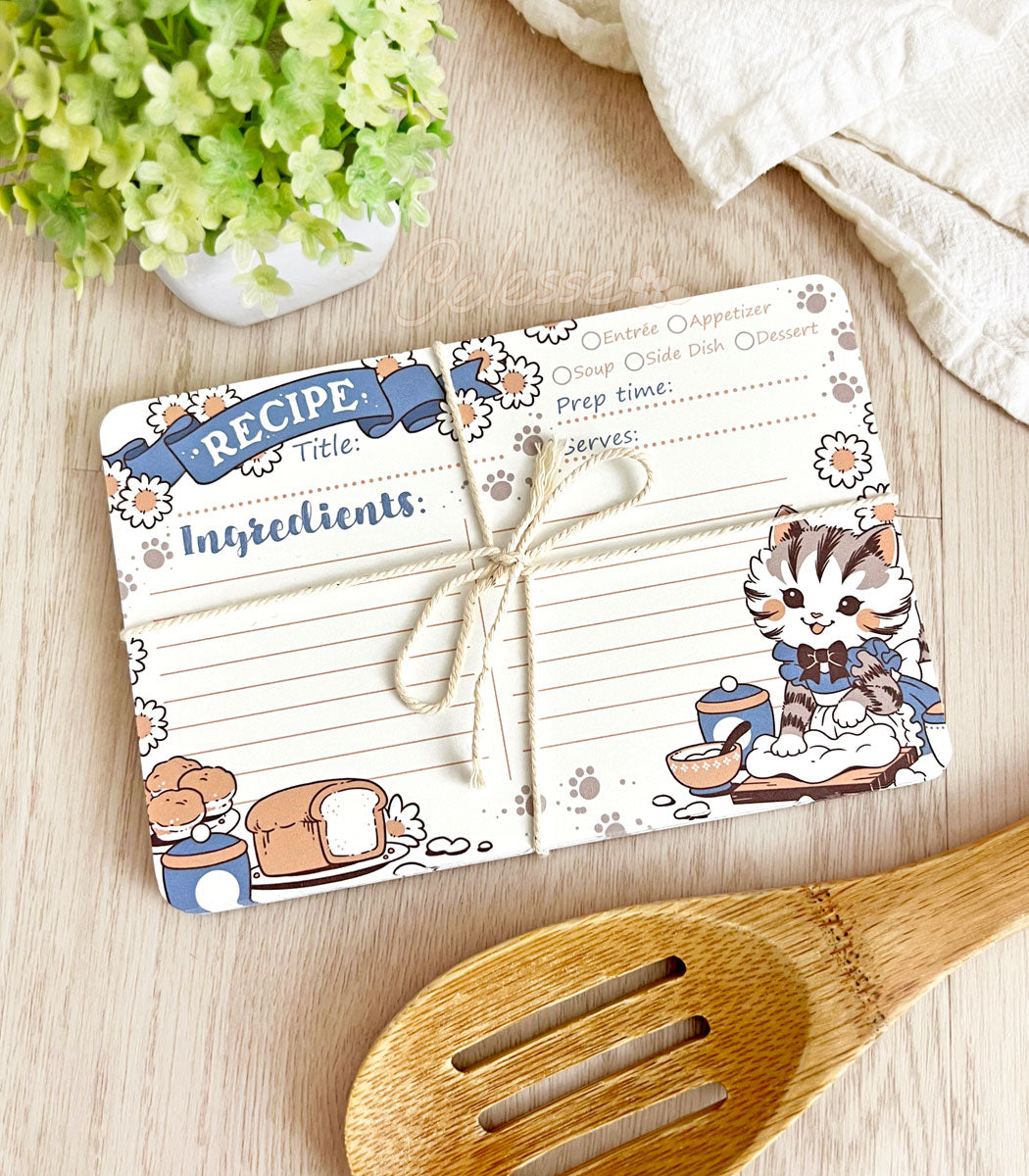 A pack of recipe cards tied up with twine on a wooden surface with kitchen utensils, the design featuring vintage-style art of a baking kitten wearing a frilly dress and apron.