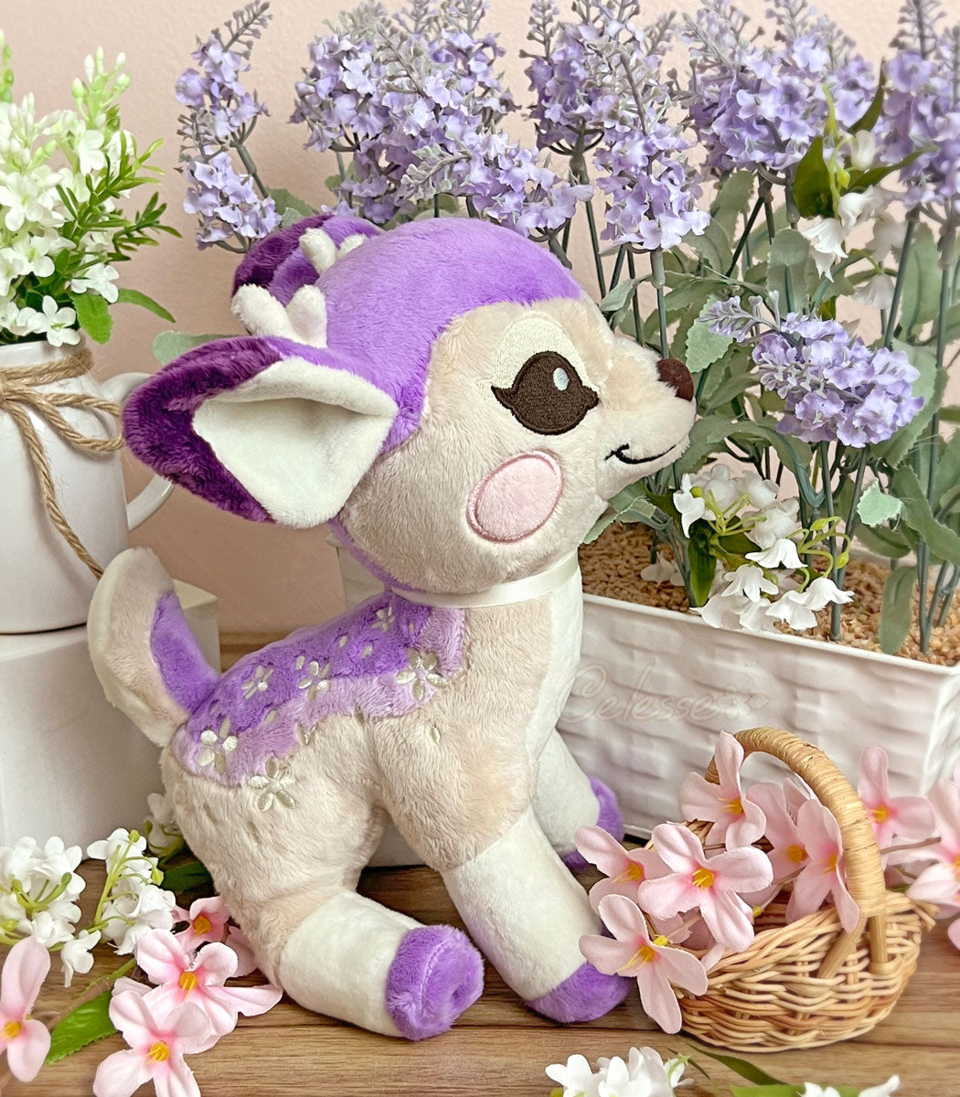 A photo of a purple baby deer plush with white flower spots posed on a table, surrounded by flowers and looking happily up at stalks of lavender.