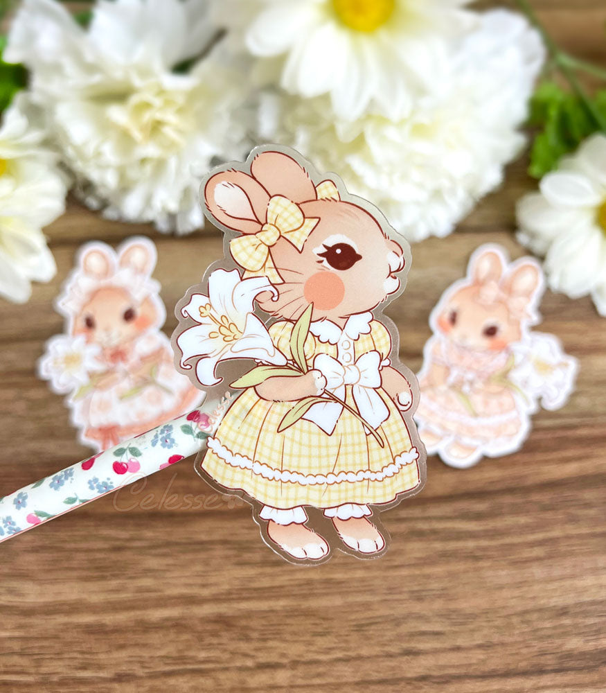 A photo of a clear vinyl sticker stuck to the end of a flowery pen, the artwork featuring a young bunny girl in a frilly vintage-style gingham dress and holding a large white lily. Two other bunny girl stickers are blurred in the background on a wood table with white flowers.