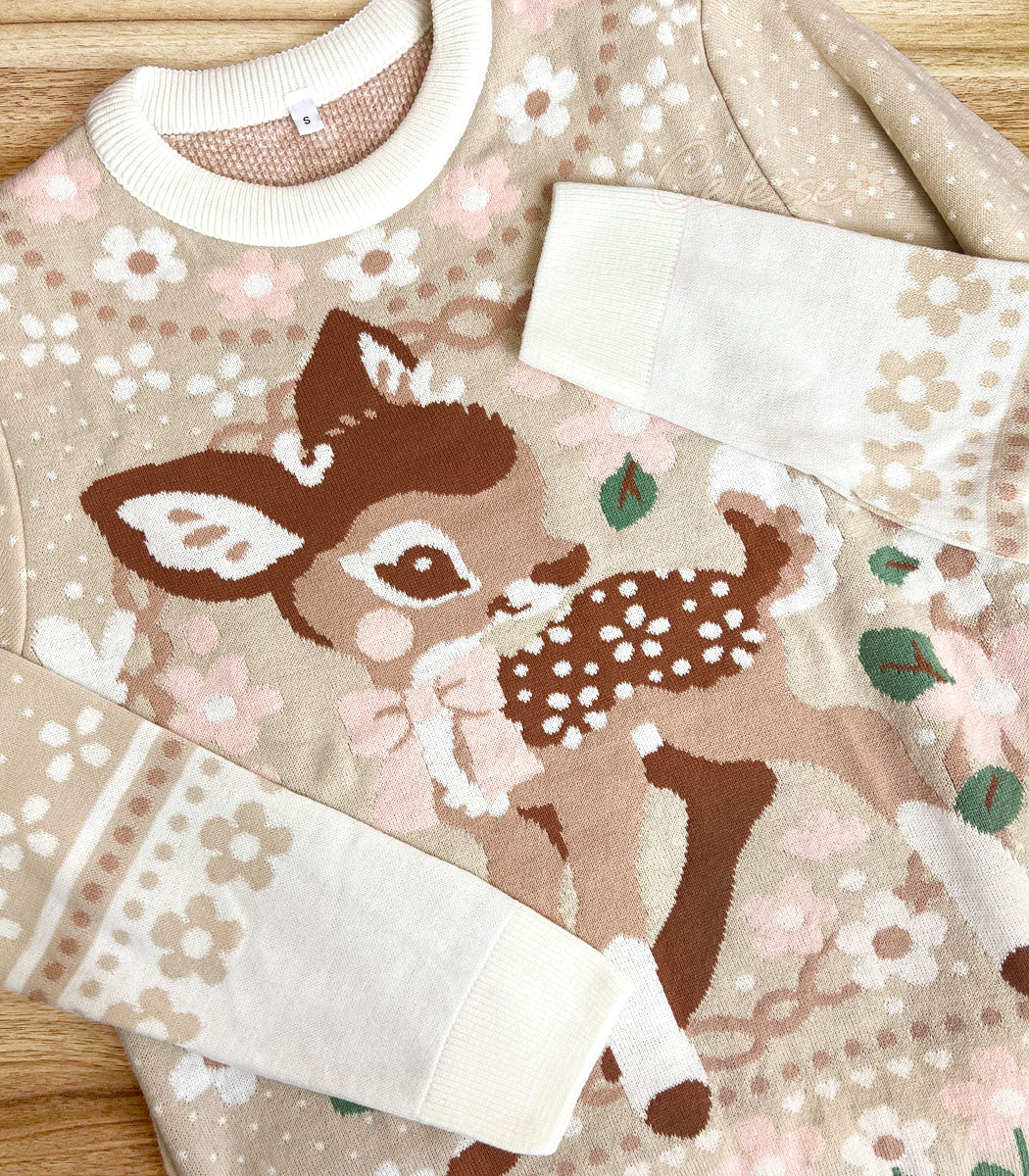A photo of a knit sweater laid on a wood table with the sleeves folded in so the cuffs lay in diagonals across the front. The colors are soft beige and browns and the front design features an image of a baby deer standing in a floral wreath.