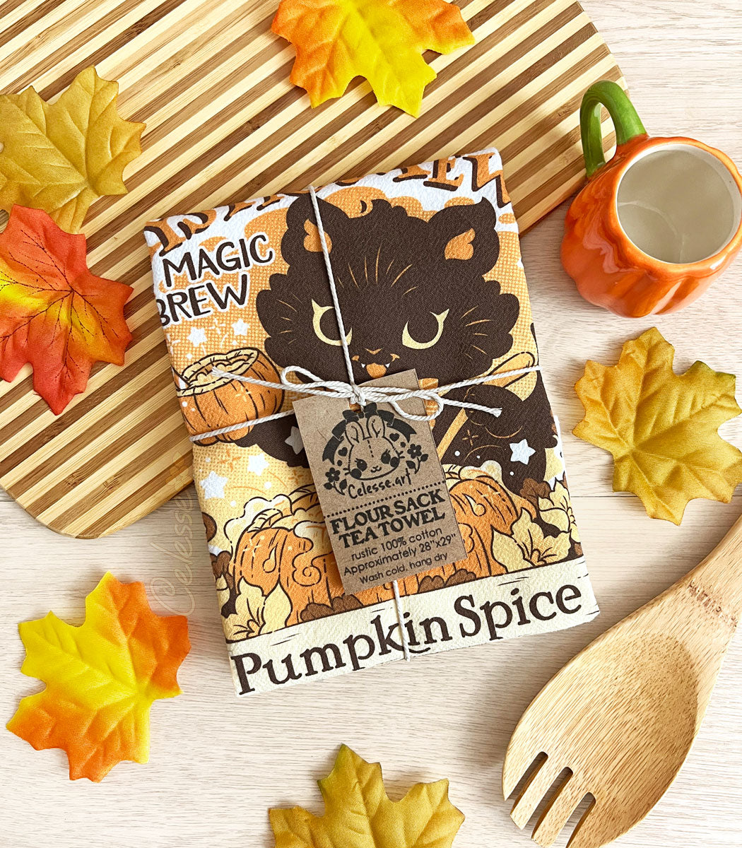 A photo of a tea towel folded into a square and tied with twine, featuring a print with a black cat stirring a pumpkin cauldron. A striped cutting board, a spoon and fall leaves decorate the background.
