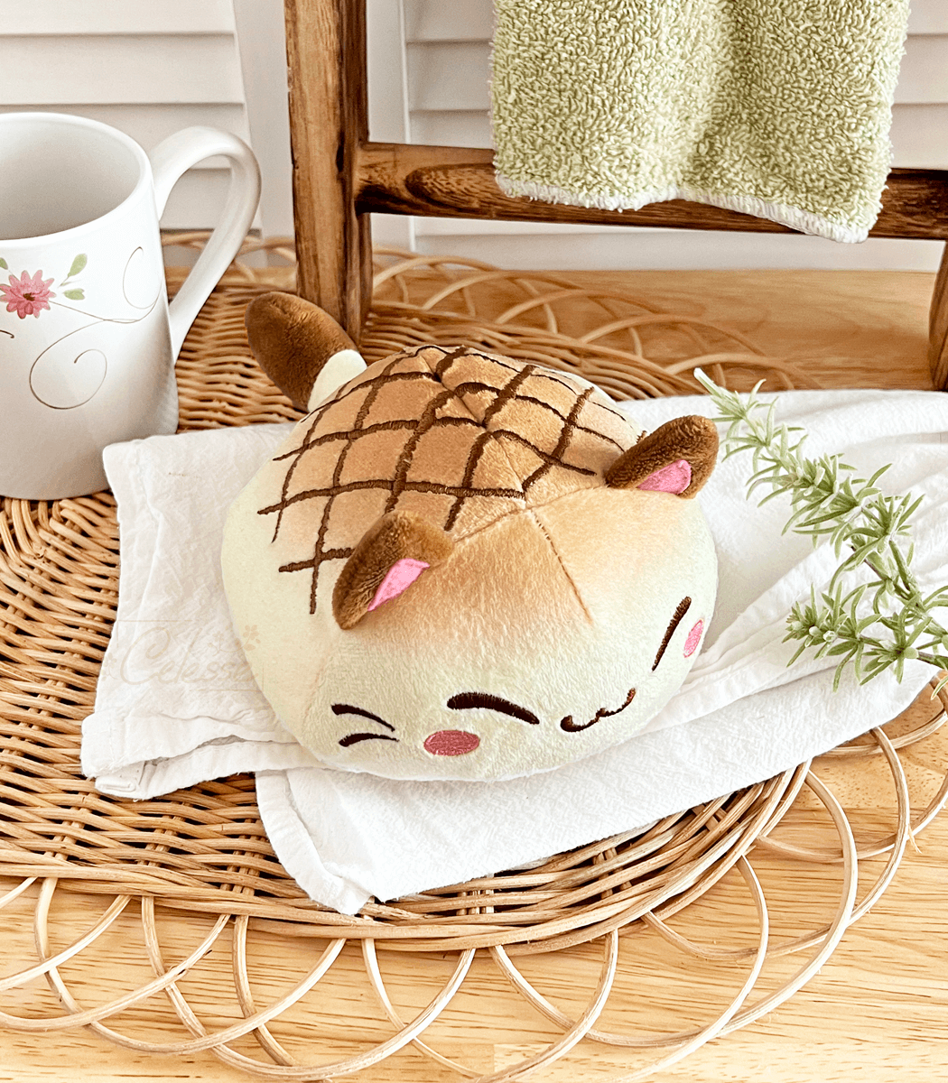 A photo of a round plush bread cat in the shape of melon bread laid out on a white towel on a kitchen counter.