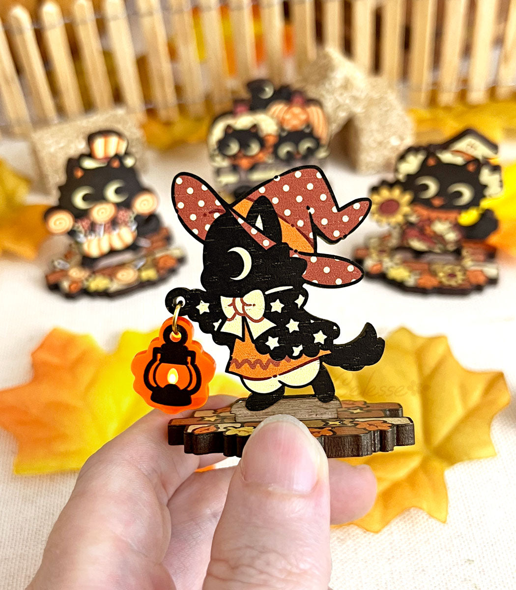 A hand holding a wooden standee depicting whimsical black cat characters in Halloween attire, arranged on a surface with autumn leaves and a picket fence.