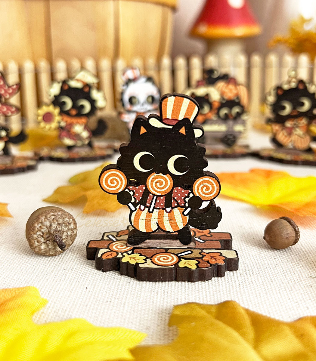 A wooden standee depicting whimsical black cat characters in Halloween attire, arranged on a surface with autumn leaves and a picket fence.