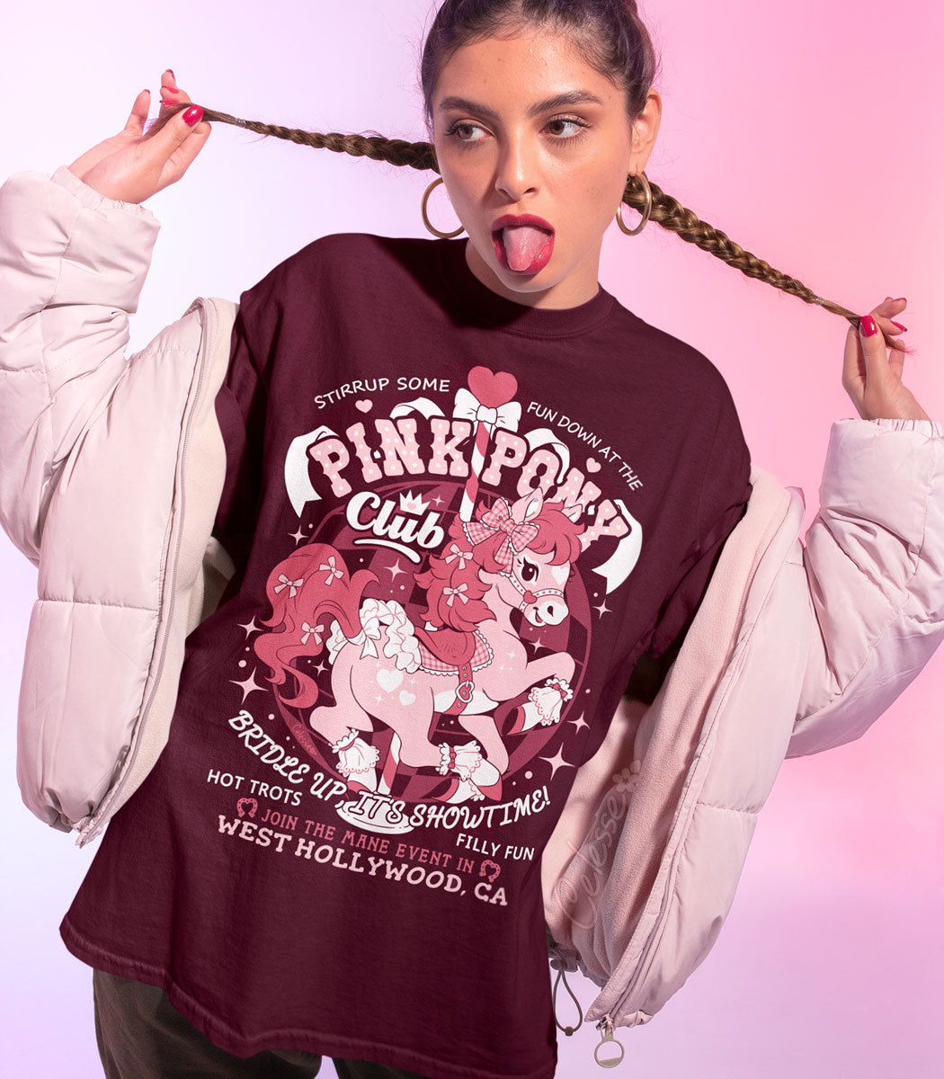 A woman modeling a dark maroon graphic t-shirt with a faux logo for the Pink Pony Club, styled with a light pink puffer jacket against a pink gradient background.