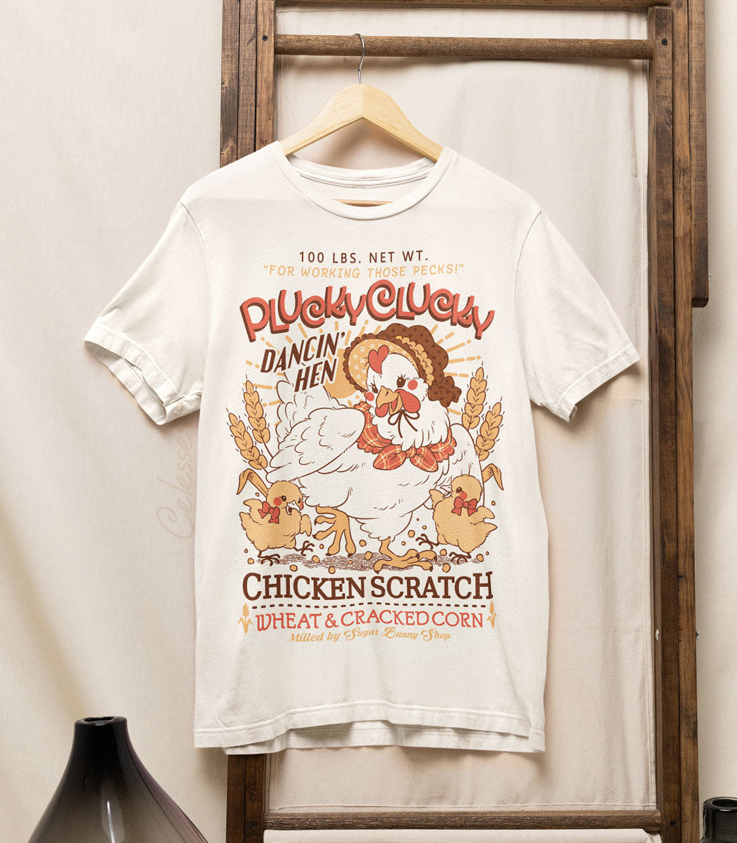A photo of an off-white shirt on a wood hanger, hung from a wooden frame, the printed design features a vintage-style logo for Plucky Clucky Dancin&