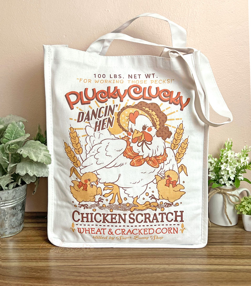 A photo of a large white tote bag sitting on a table with plants on either side. The front print features a faux vintage-style logo for Plucky Clucky Chicken Scratch with a dancing hen in a bonnet and two chicks.