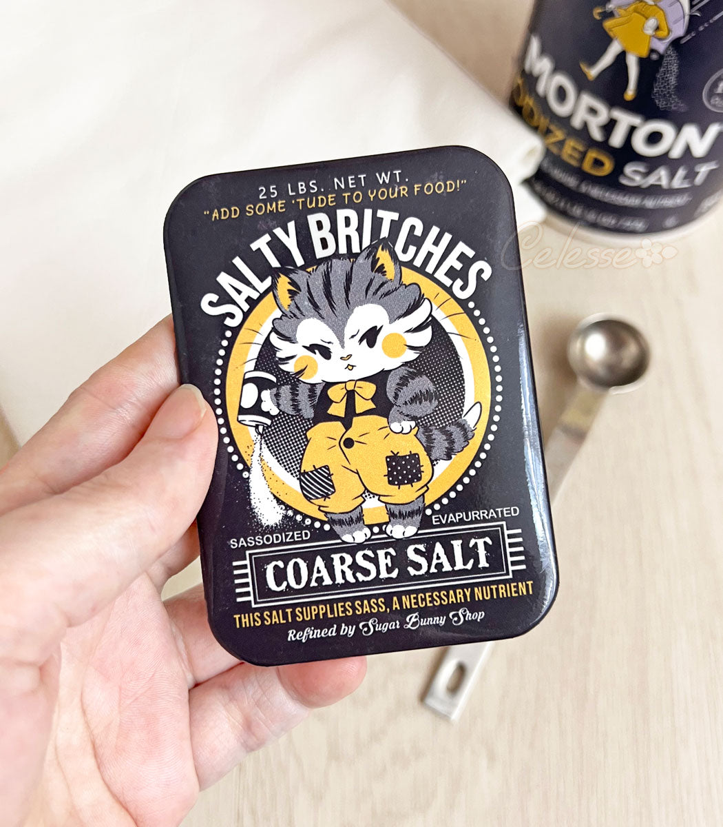 A photo of a hand holding a fridge magnet with artwork featuring a vintage-style logo for Salty Britches Coarse Salt with a kitten wearing patchwork britches and petulantly pouring out a container of salt.